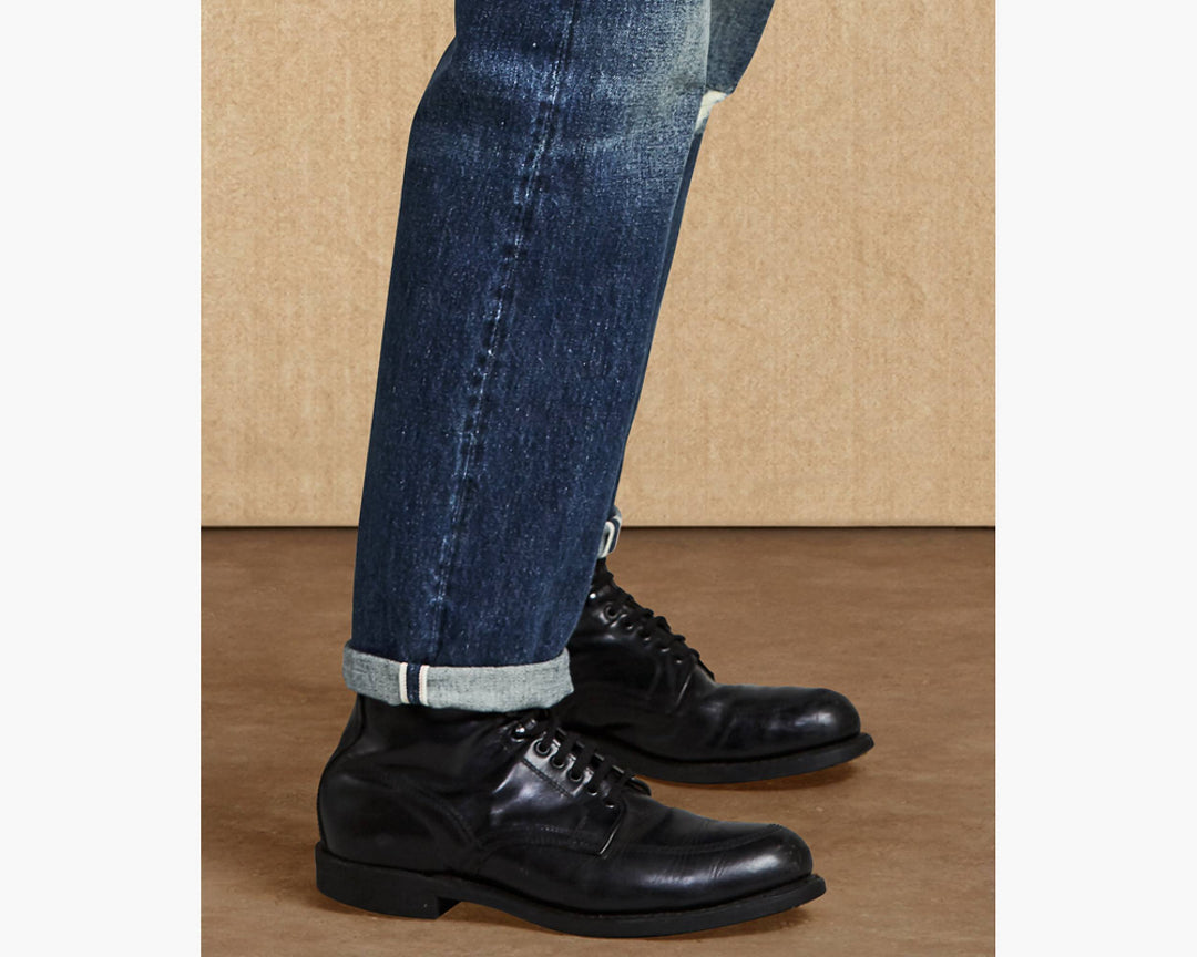 Black leather shoes and blue jeans on a brown floor.