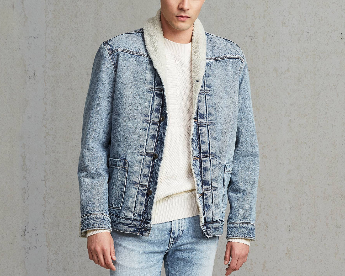 Man wearing a denim jacket over a white sweater with light blue jeans against a gray background