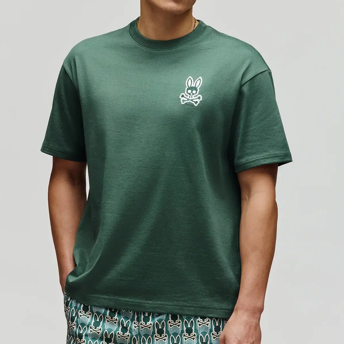 Man wearing a green t-shirt with a logo and patterned shorts on a light gray background